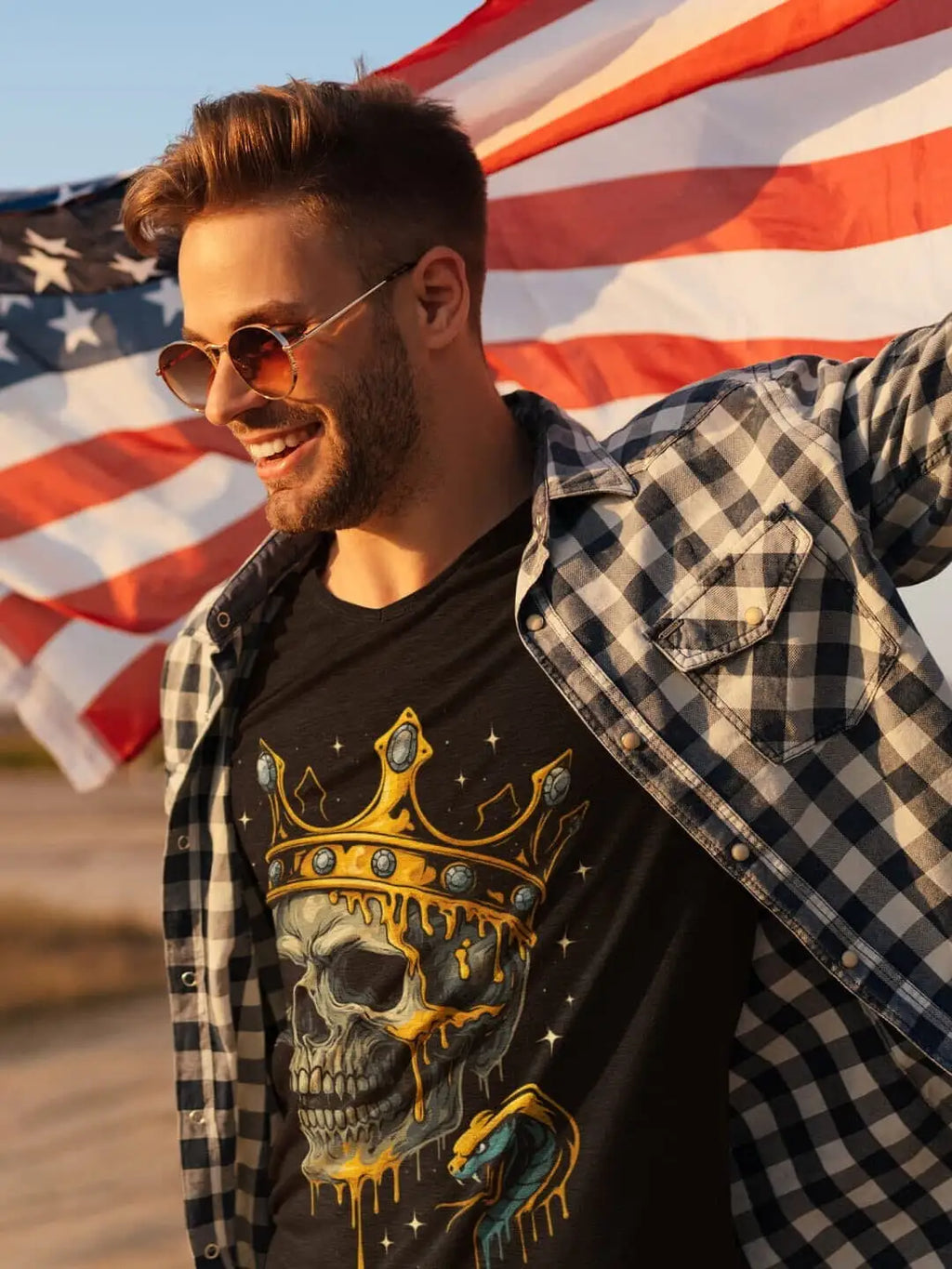 A smiling man wearing a black t-shirt with a skull, crown, and snake design, under an open plaid shirt, with an American flag in the background