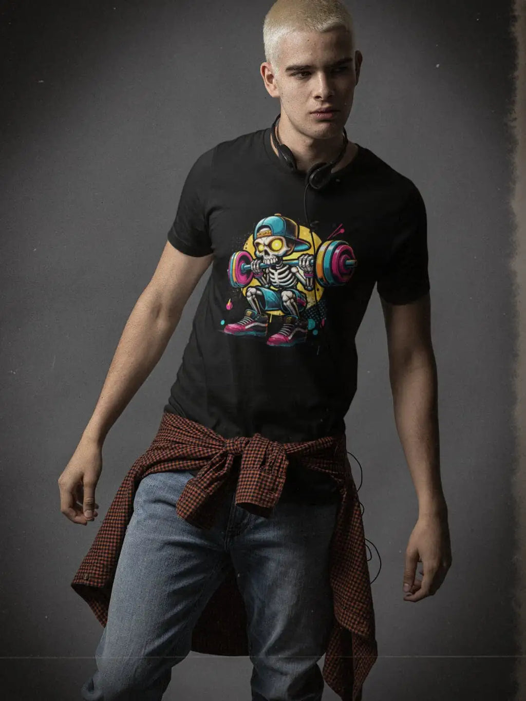 A young man with platinum blonde hair wearing a black t-shirt with a colorful skeleton graphic, light-wash jeans, and a red and black plaid shirt tied around his waist
